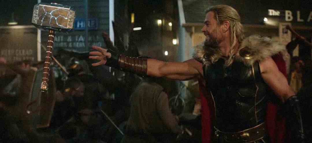 Thor, dressed in armor and a red cape, extends his arm to summon his hammer, Mjolnir, amid a chaotic battle scene with blurred figures and city lights in the background.