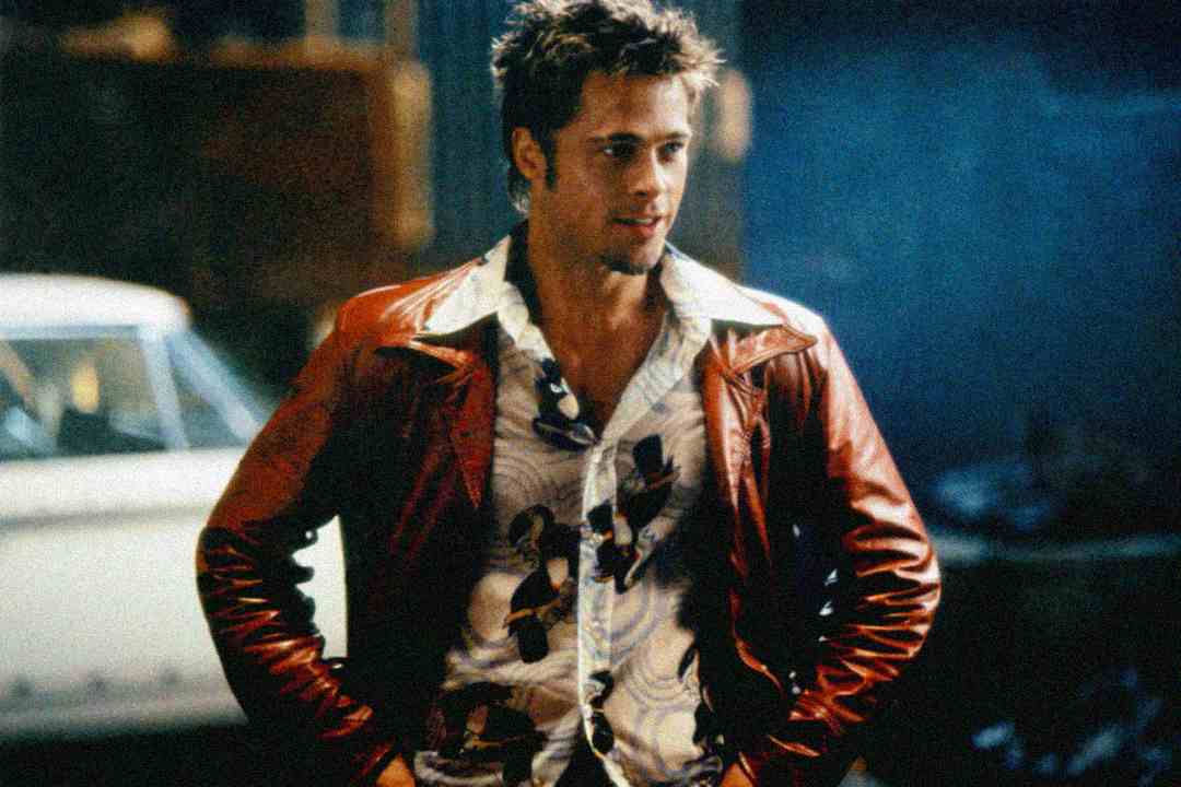 A man with spiked hair wears a red leather jacket over a patterned shirt, standing with his hands on his hips. A vintage white car is blurred in the background. The scene has a gritty, urban feel.