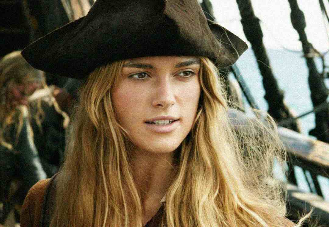 A woman with long blonde hair wearing a large black pirate hat stands on a wooden ship, with rigging and sails visible in the background. She looks ahead with a calm, determined expression.