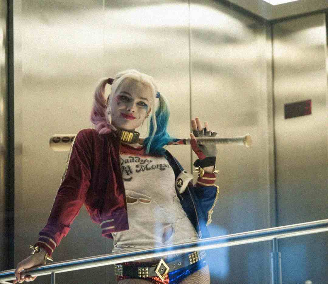 A person in a Harley Quinn costume with pink and blue pigtails, a baseball bat over their shoulder, and wearing a red jacket and "Daddy's Lil Monster" shirt stands in an elevator, smiling confidently.