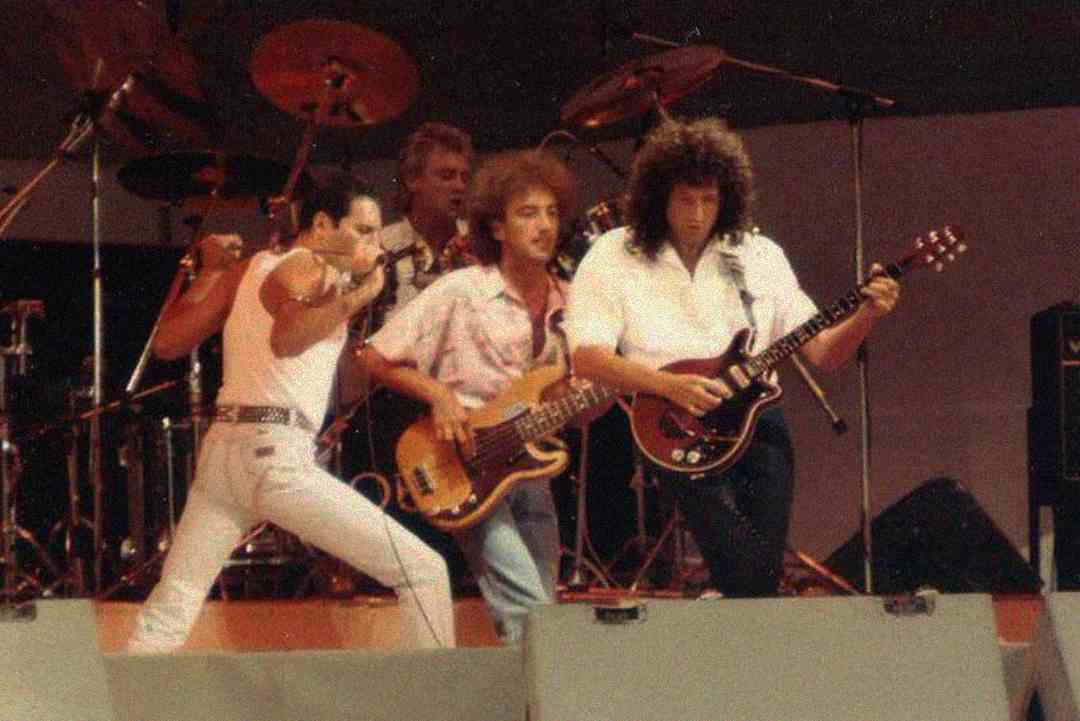 Four musicians perform energetically on stage with instruments and microphones, including a guitarist, bassist, drummer, and lead vocalist, under stage lighting and surrounded by drum equipment.