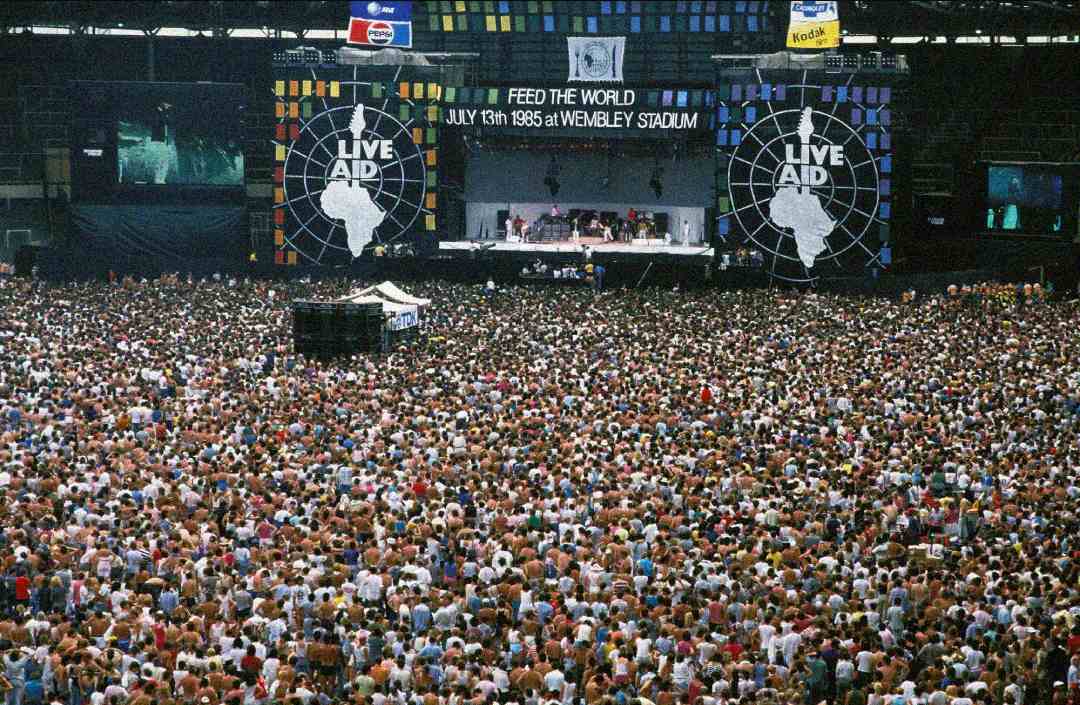 A massive crowd fills Wembley Stadium for the 1985 Live Aid concert. The stage features "LIVE AID" and “FEED THE WORLD JULY 13th 1985 AT WEMBLEY STADIUM” banners, with musicians performing and large screens overhead.