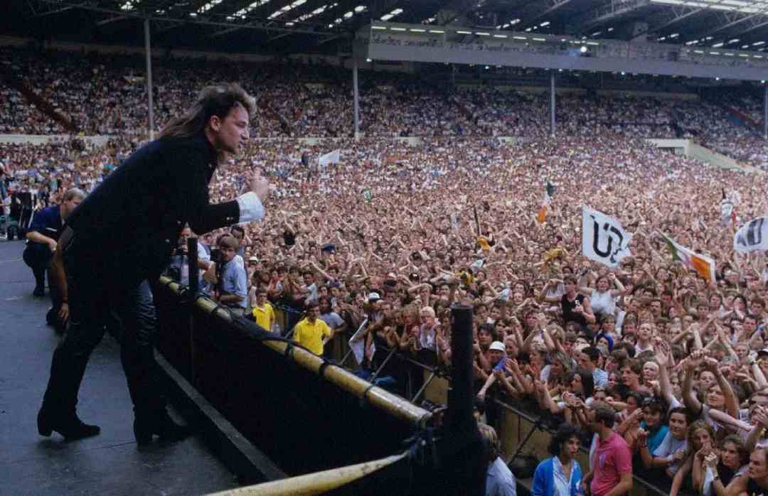 A performer stands at the edge of a stage, leaning towards a huge crowd of cheering fans in a packed stadium, with flags waving and people raising their hands in excitement.