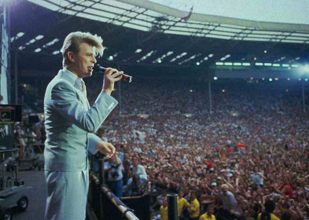 A singer in a light blue suit performs on stage at a large stadium, holding a microphone, with a massive crowd of cheering fans filling the stands and floor area.