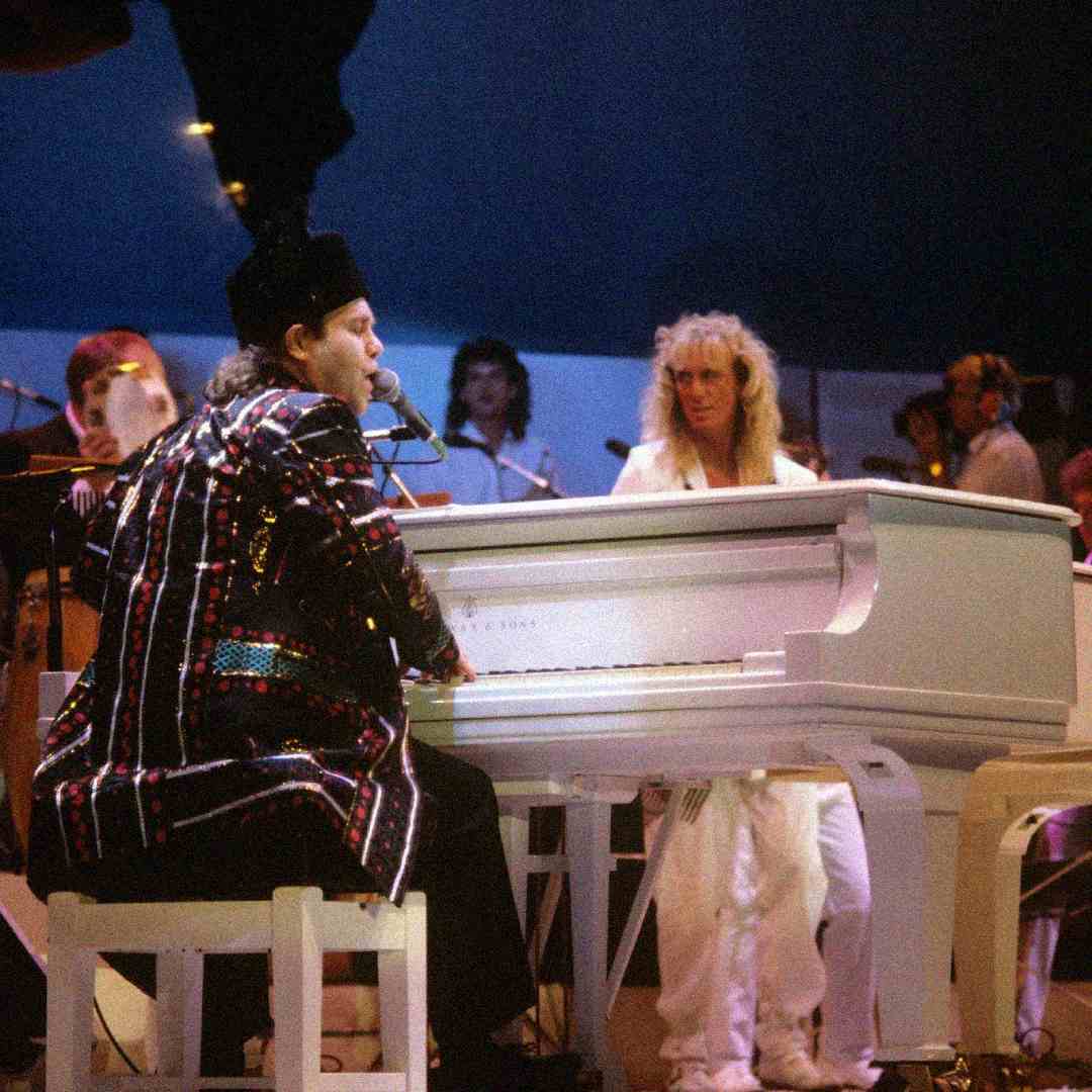 A musician wearing a colorful, sequined outfit plays a white grand piano on stage, accompanied by other performers, including one with long blond hair dressed in white, under dramatic stage lighting.