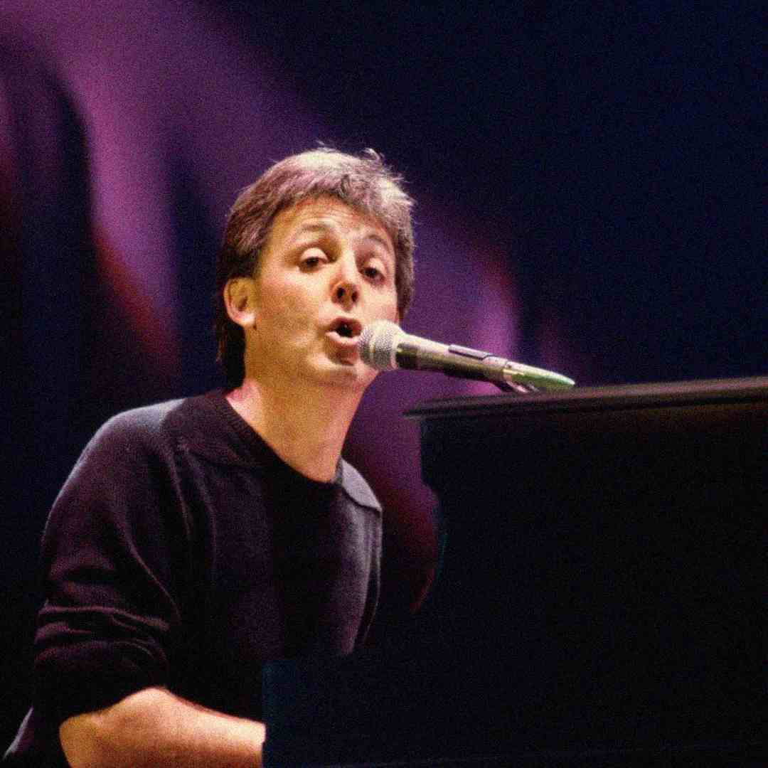 A person with short brown hair sings into a microphone while playing a piano on stage, wearing a black shirt, with a blurred, colorful background.