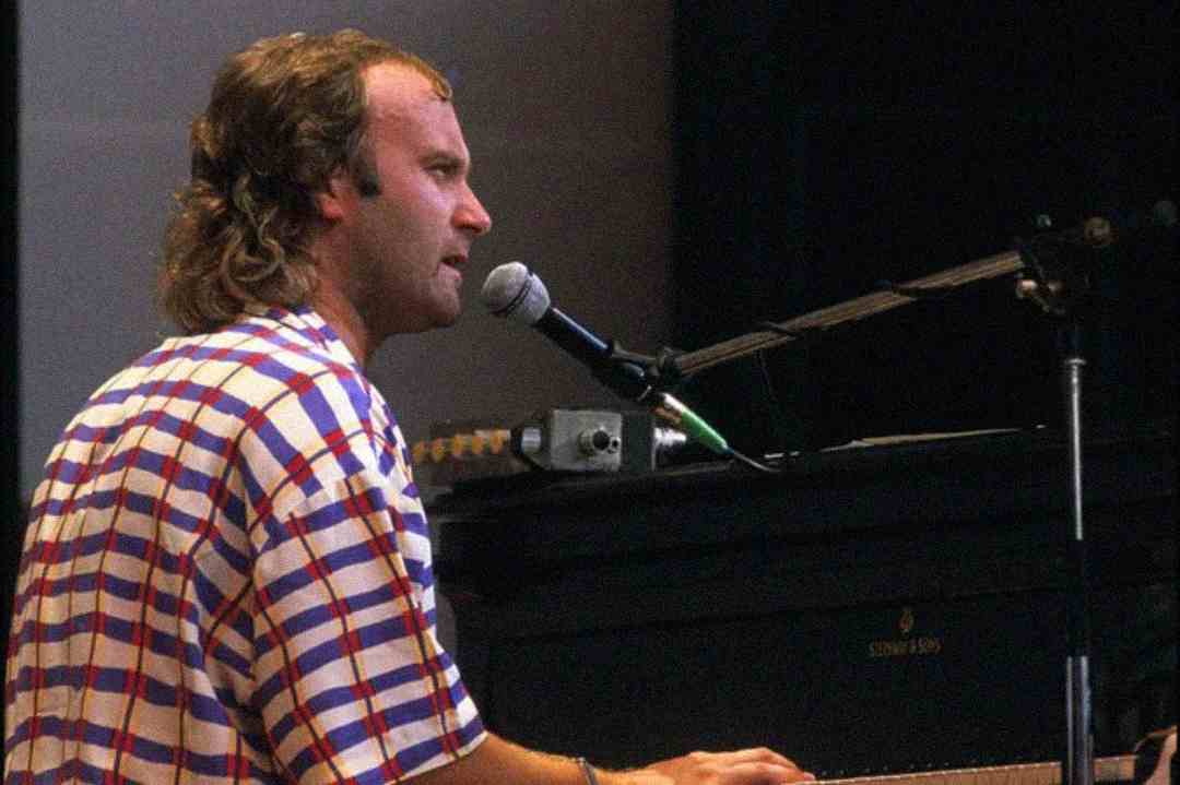 A man with short, curly hair and a mullet, wearing a red, blue, and white plaid shirt, sings into a microphone while playing a black grand piano on stage.