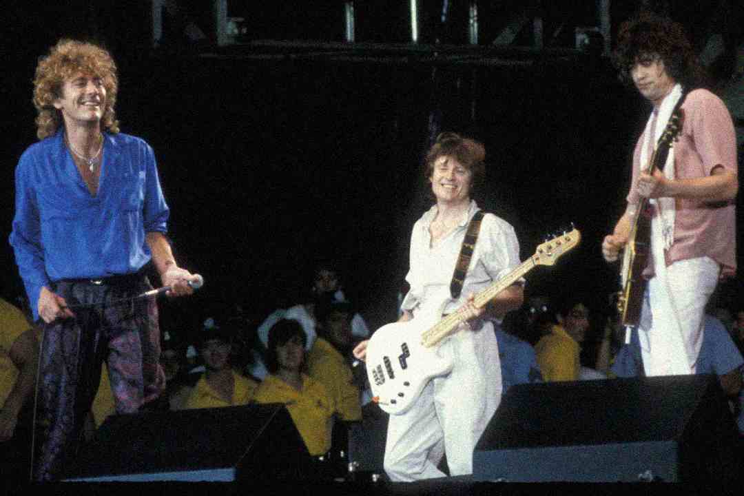 Three musicians perform on stage; one sings holding a microphone, another plays a white guitar, and a third plays an electric guitar. They are smiling; security personnel in yellow shirts stand in the background.