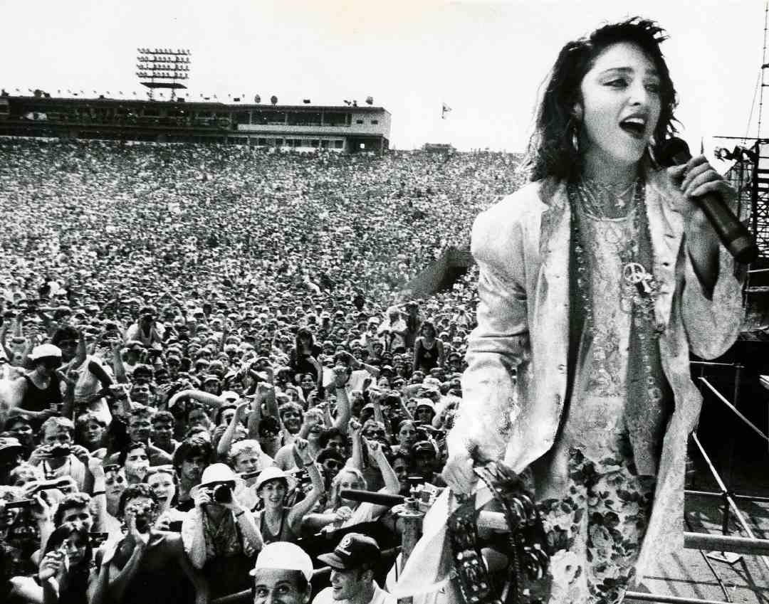 A woman sings into a microphone on stage in front of a massive outdoor crowd. She wears layered clothing and jewelry. The cheering audience fills the stadium behind her. The image is in black and white.