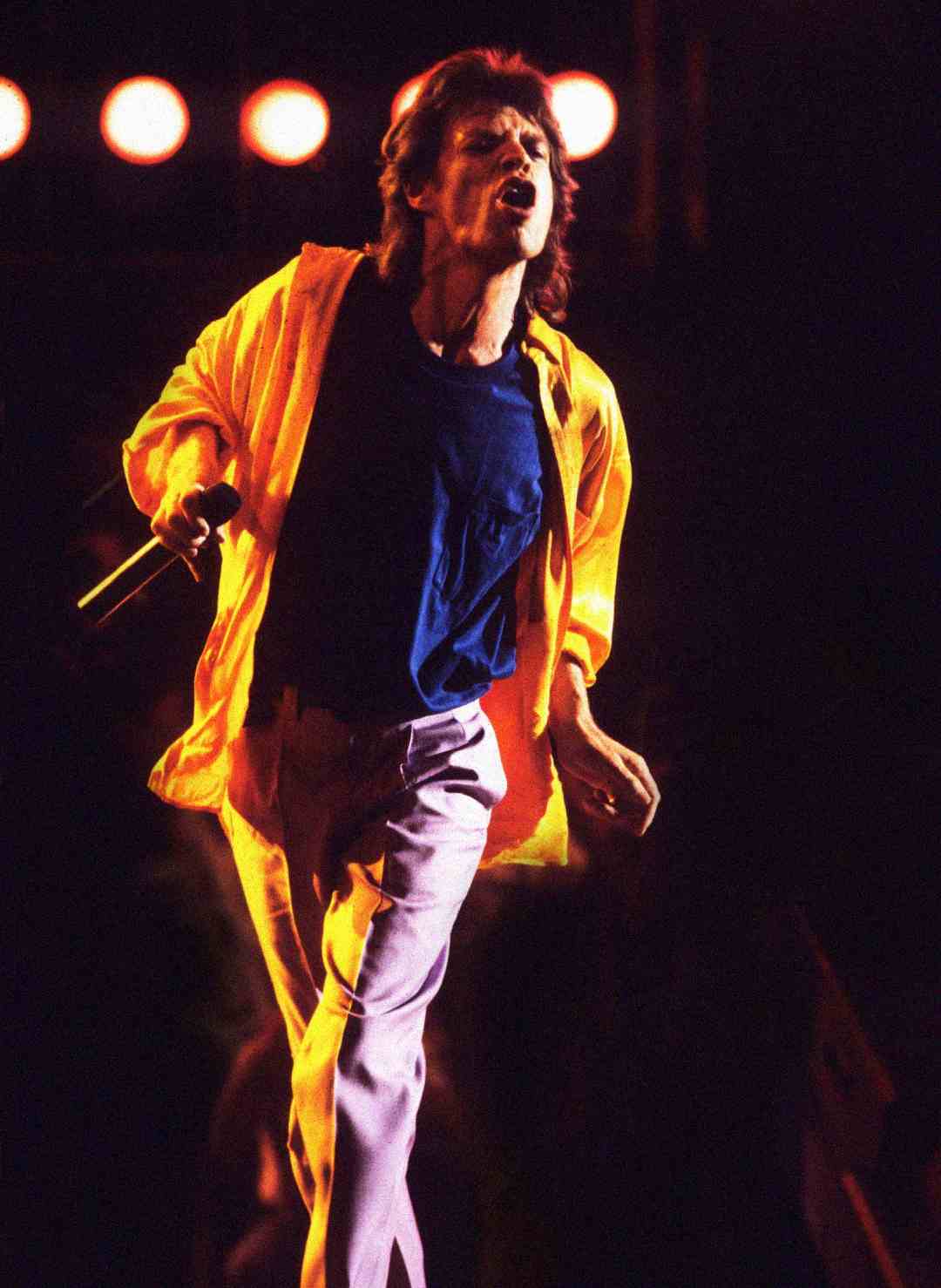 A person with long hair, wearing a yellow jacket and blue shirt, energetically sings into a microphone on stage under bright lights.