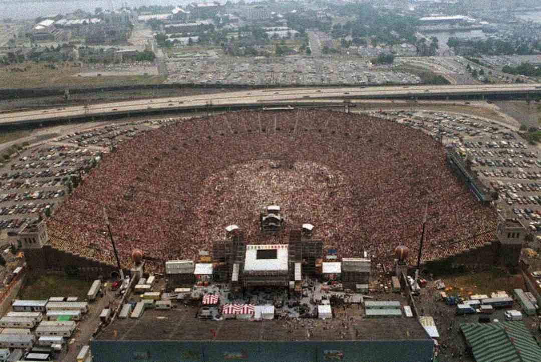 A large stadium filled with thousands of people attending an outdoor concert, with a stage set up in the center and parking lots and highways visible in the background.