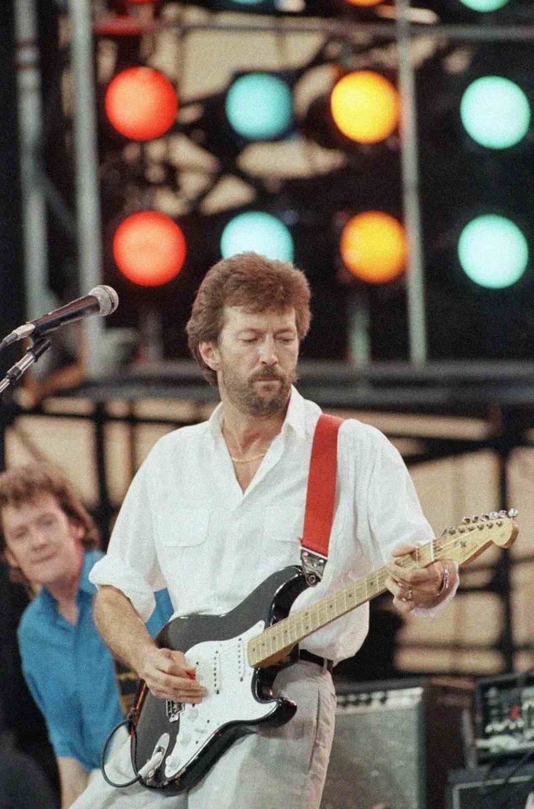 A man with a beard wearing a white shirt plays an electric guitar on stage, with colorful stage lights glowing in the background and another musician visible behind him.
