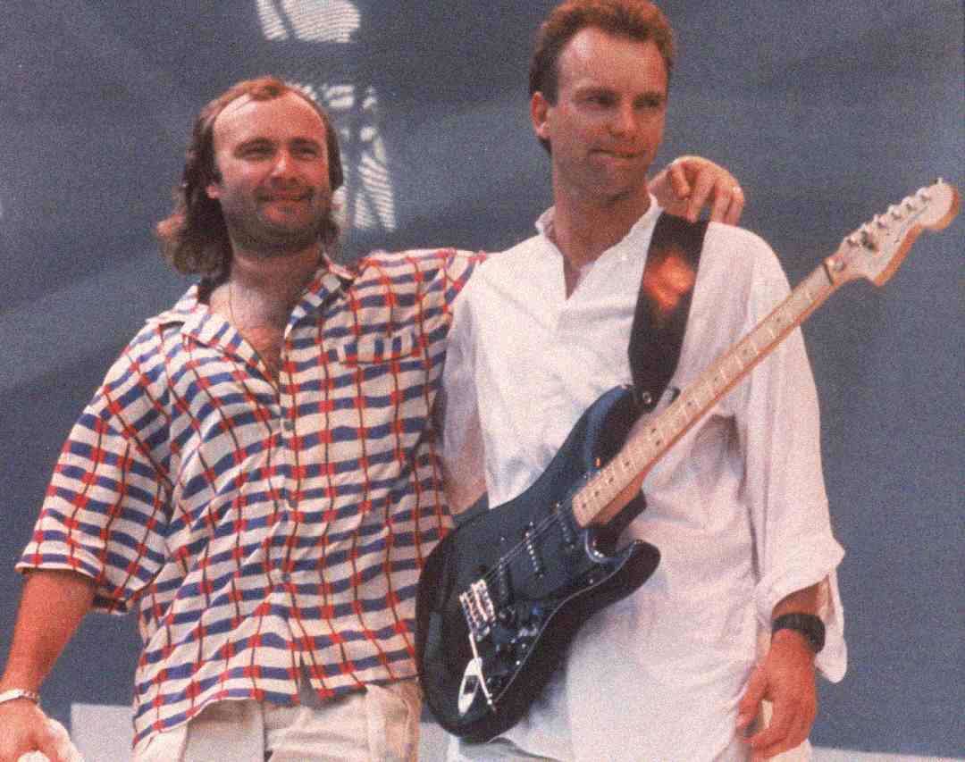 Two men stand together on stage; one has his arm around the other. The man on the left wears a checked shirt, while the man on the right, dressed in a white shirt, holds a black electric guitar. Both are smiling.