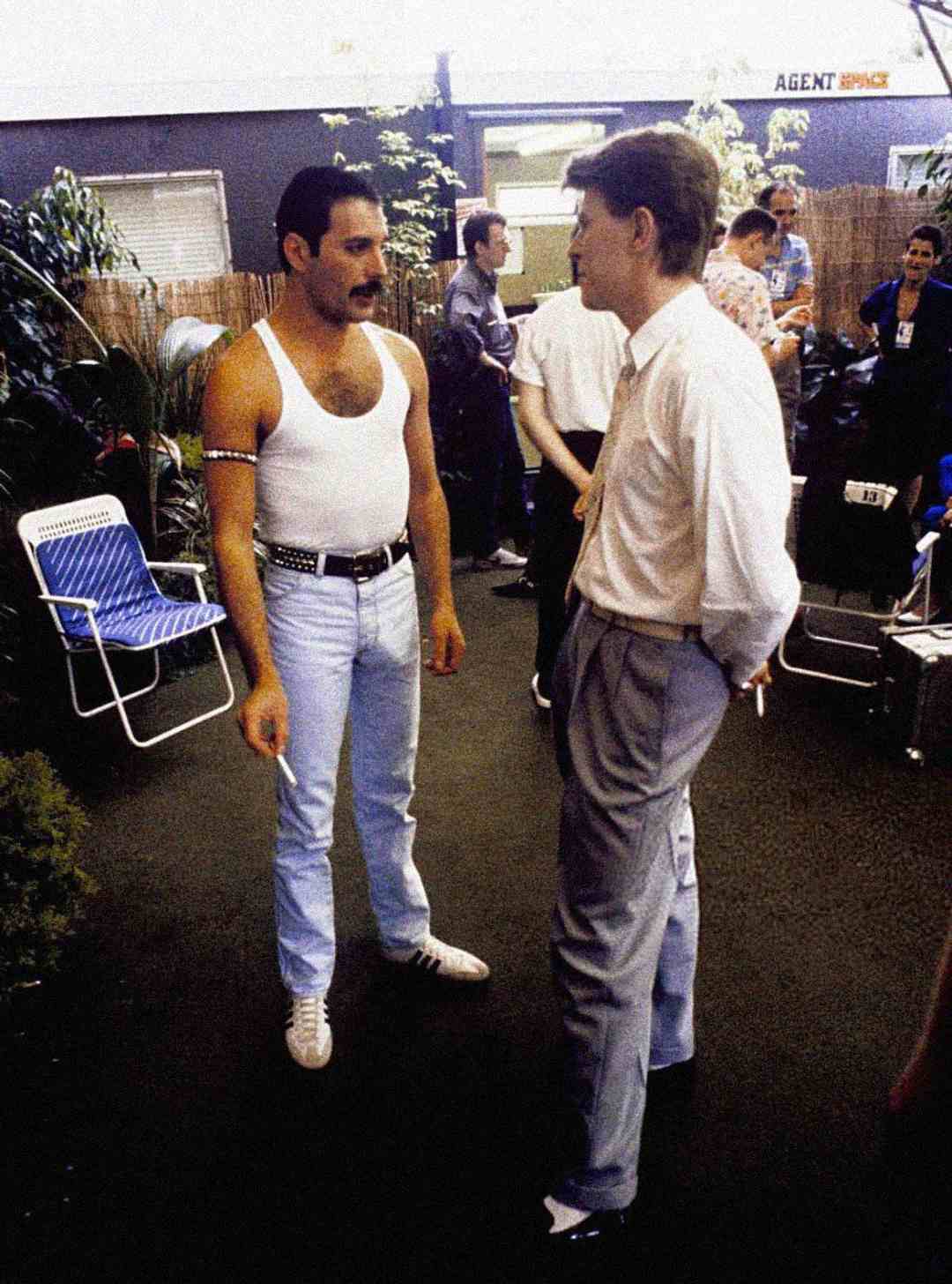A man in a white tank top and light blue jeans talks to another man in a white shirt and grey pants in a backstage area, with other people and equipment visible in the background.