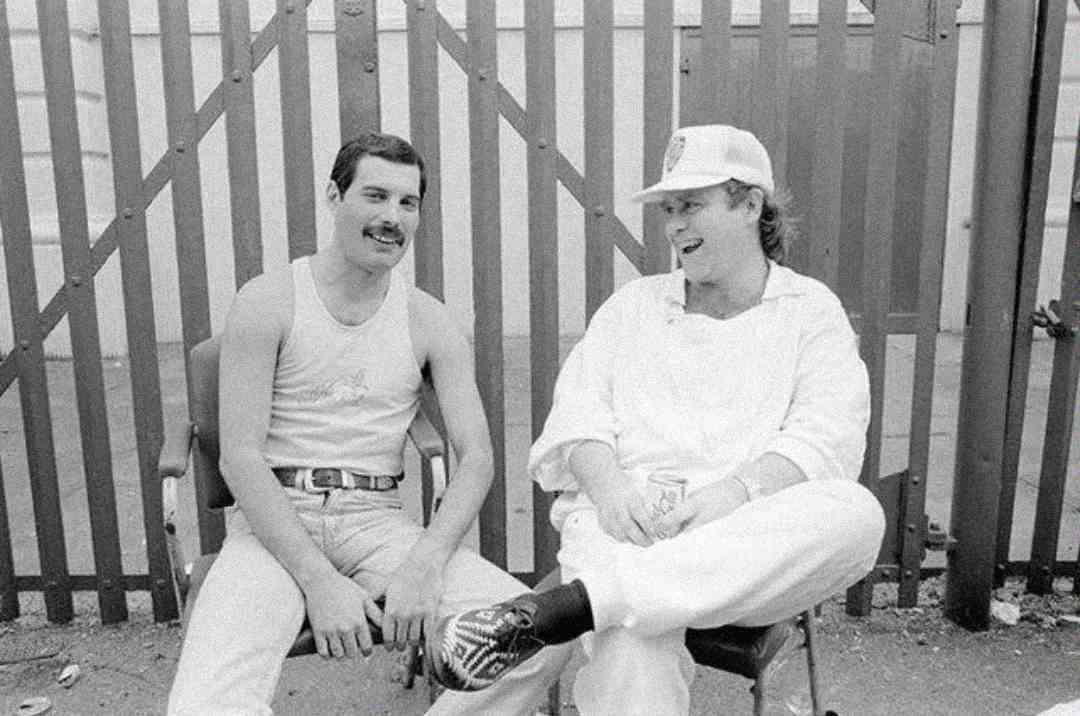 Two men sit on chairs outdoors, smiling and talking. One wears a tank top and jeans, the other wears white clothes and a hat. A metal fence and building are in the background.
