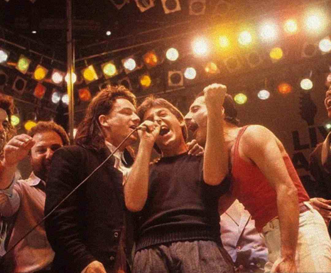 A group of musicians passionately sing together on stage under bright lights, surrounded by an enthusiastic crowd, capturing the energy of a live concert performance.