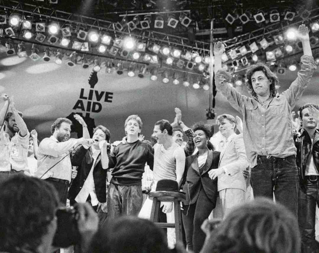 A group of musicians and performers stand on stage under bright lights, some with raised arms, during the Live Aid concert. The "LIVE AID" logo is visible in the background. The crowd is visible in the foreground.