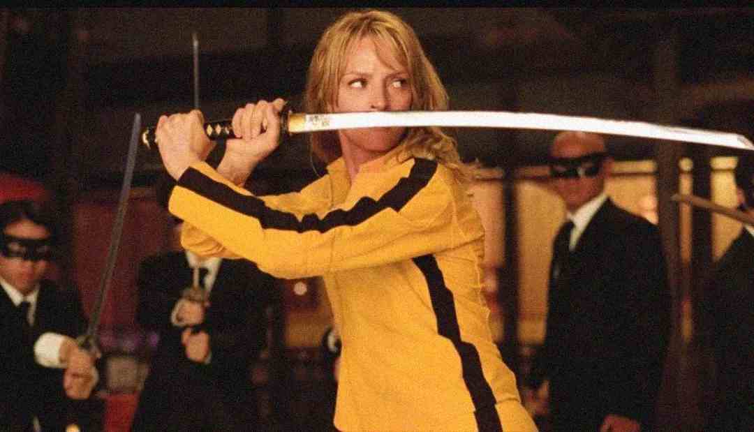 A woman in a yellow suit holds a katana in a defensive stance, surrounded by people in black suits and masks in a dimly lit room.