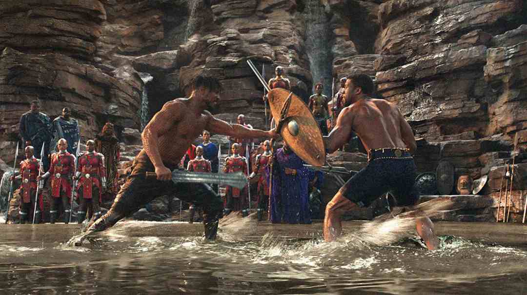 Two shirtless men duel with weapons and shields in shallow water, surrounded by rocky cliffs and onlookers in red and blue attire, creating an intense and dramatic scene.