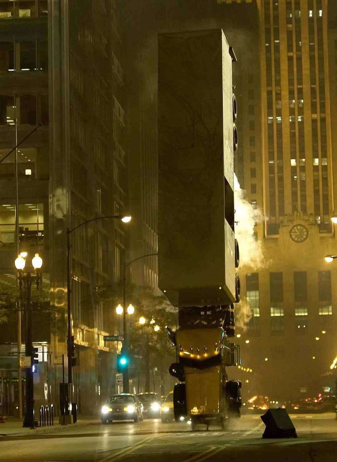 A large truck stands vertically on its rear end in the middle of a city street at night, with smoke rising around it and illuminated buildings in the background.