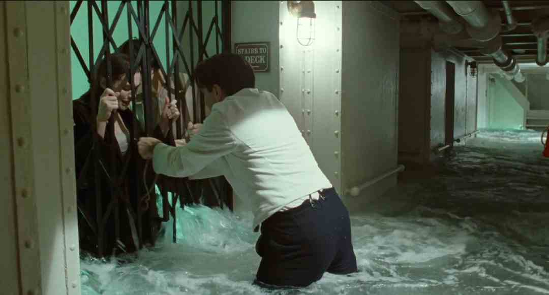 A man stands in waist-deep water, trying to open a metal gate to rescue a woman trapped behind it in a flooded, dimly lit corridor with pipes and a “Stairs to Deck” sign on the wall.