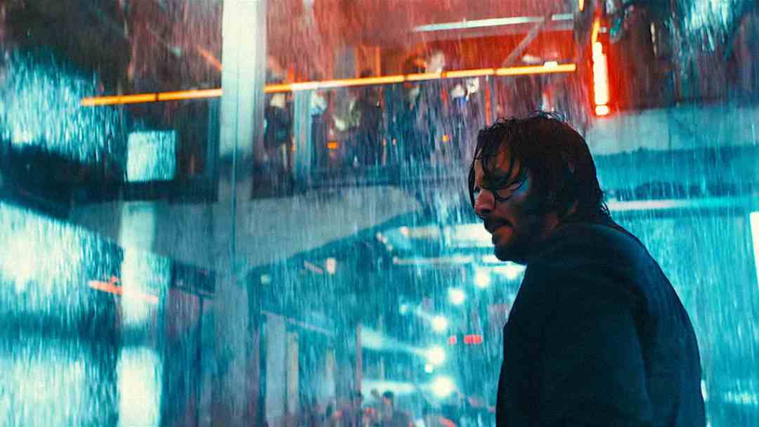 A man with wet hair and beard stands under heavy rain inside a building with neon lights, looking over his shoulder. The scene is dramatic and intense, with blurred figures in the background.