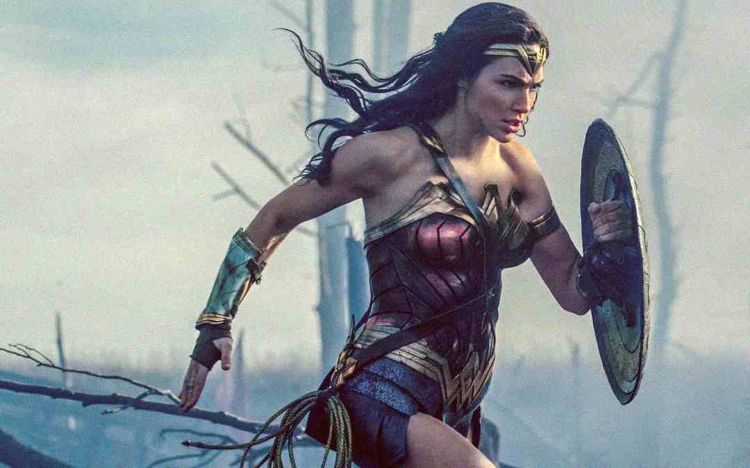 A woman in warrior armor, holding a shield and running forward in a battle scene. She has long dark hair, a metal headband, and determined expression, with a smoky, war-torn background.