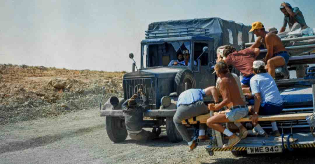 A group of people in casual summer clothing clings to the back and sides of a moving open truck on a dusty road, with someone sitting on the front bumper and another driving. The landscape is dry and barren.