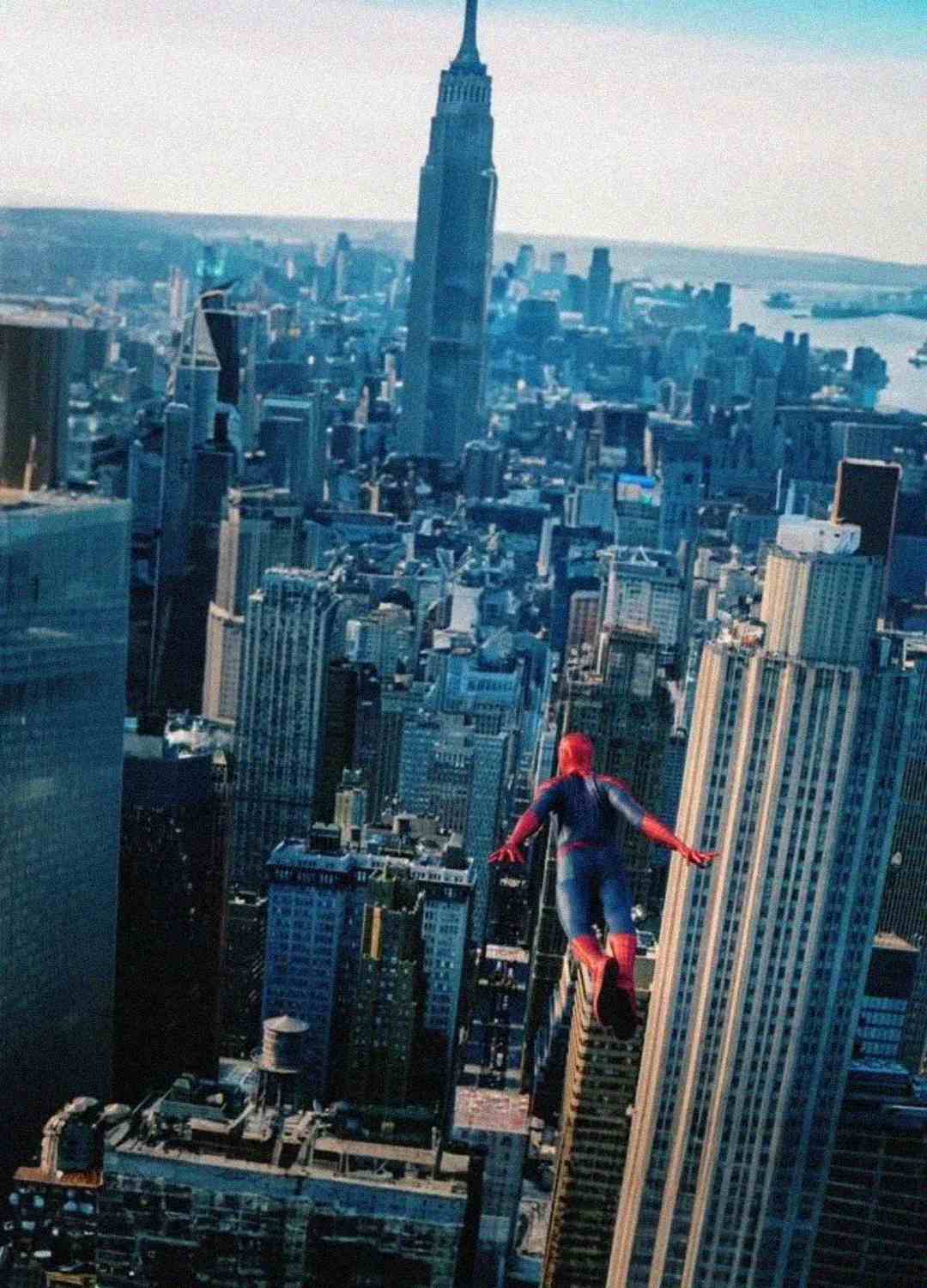 Spider-Man stands on the edge of a tall skyscraper, overlooking the New York City skyline with the Empire State Building visible in the background on a clear day.