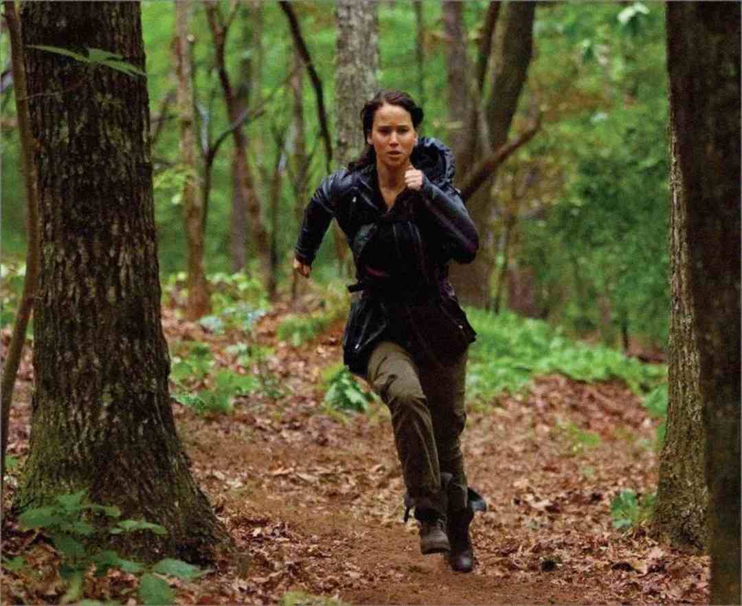 A person wearing a dark jacket runs quickly through a dense, green forest on a dirt path, surrounded by tall trees and leafy foliage.