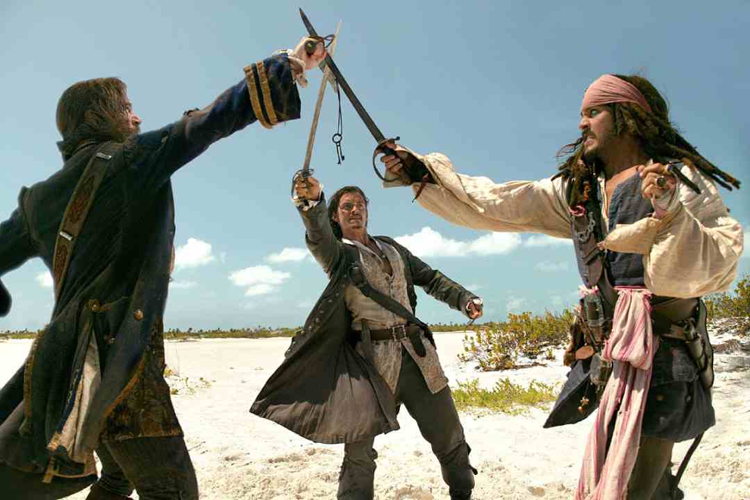 Three men dressed as pirates are engaged in a sword fight on a sandy beach under a bright blue sky, with vegetation and the ocean visible in the background.
