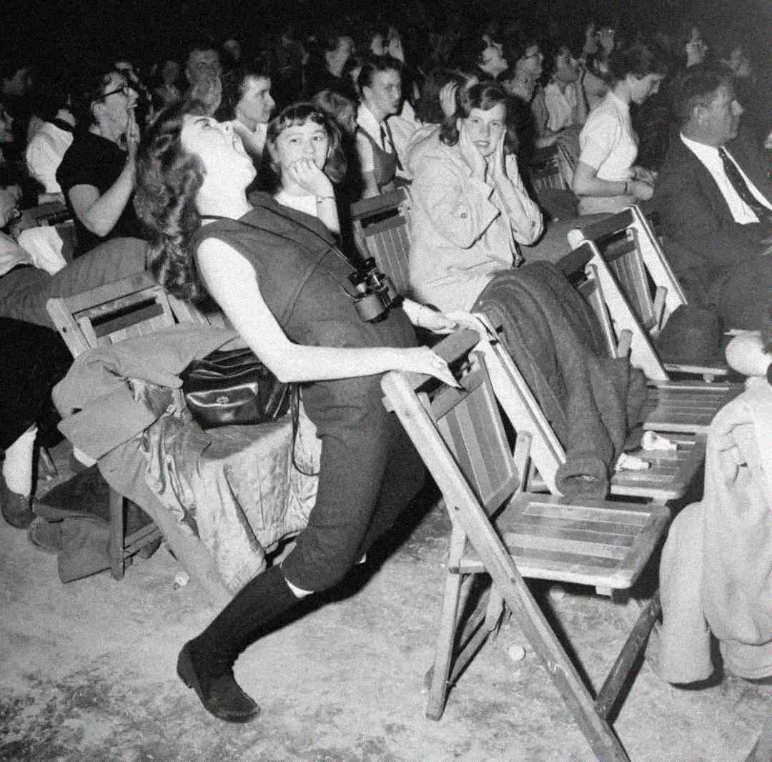 A young woman leans back dramatically in her seat, clutching the back of a folding chair at a crowded event. Others around her watch with amused or surprised expressions. The image is in black and white.