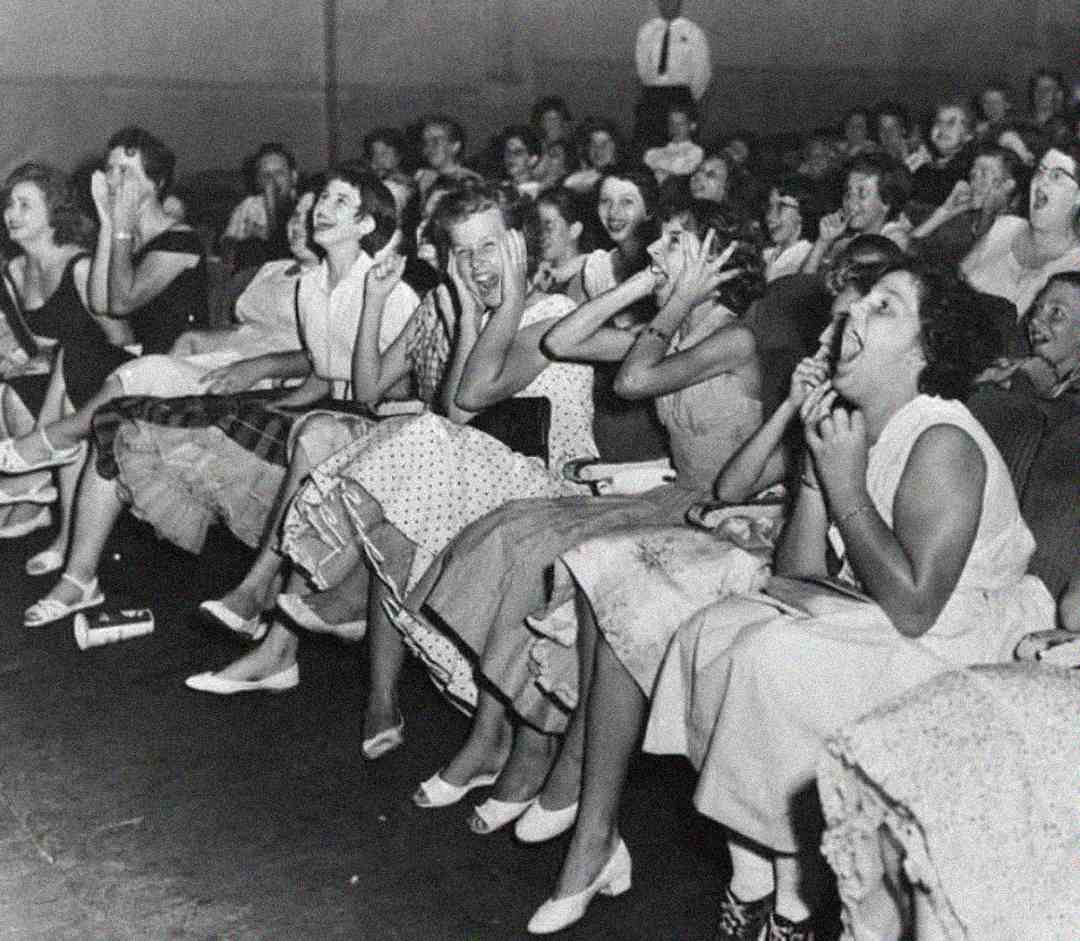 A black-and-white photo of a group of young women sitting in an audience, excitedly screaming and covering their faces with their hands. The packed crowd appears animated and thrilled, with a man standing in the background.