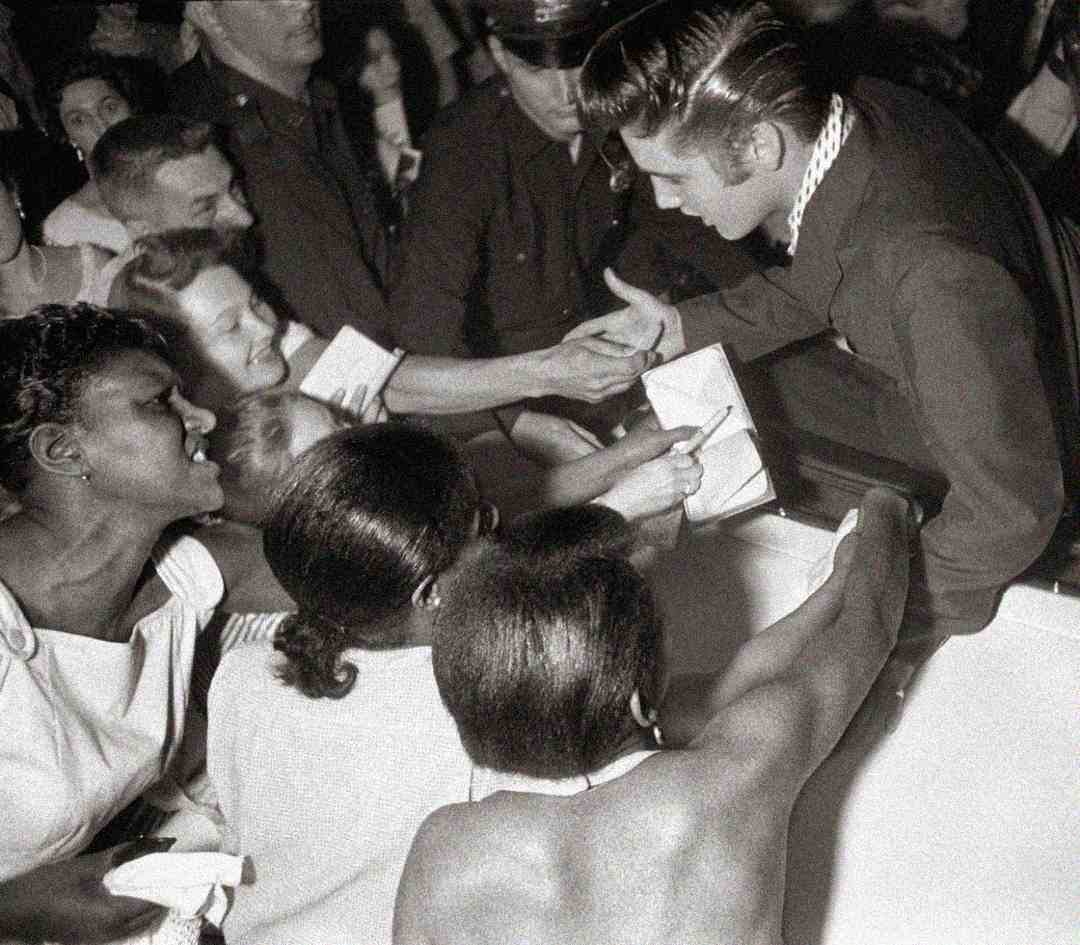 A man in a suit leans over a barrier to greet enthusiastic fans, shaking hands and signing autographs as people reach out to him and smile. A police officer stands in the background among the crowd.
