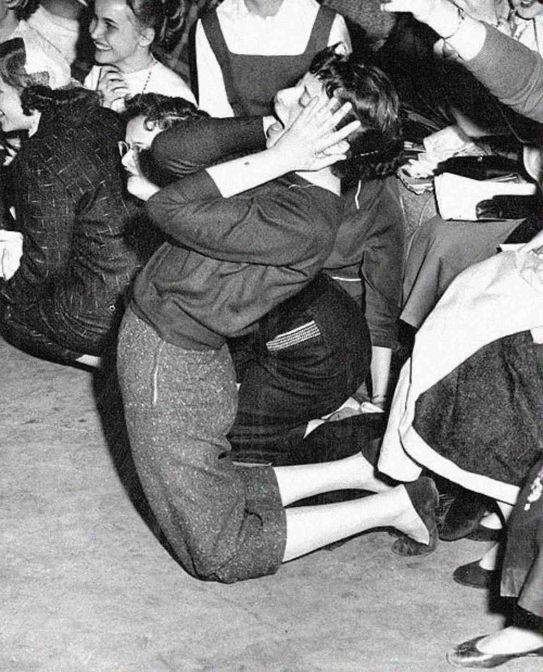 A young woman kneels on the floor, clutching her head in excitement or shock, surrounded by a crowd of other enthusiastic people at what appears to be a crowded event.