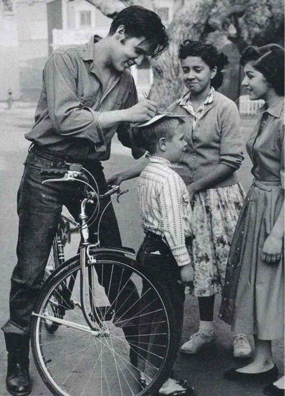 A young man signs a boy's hat while standing next to a bicycle. Two smiling girls watch the scene, and all four people are outdoors on a street. The photo is black and white and appears vintage.