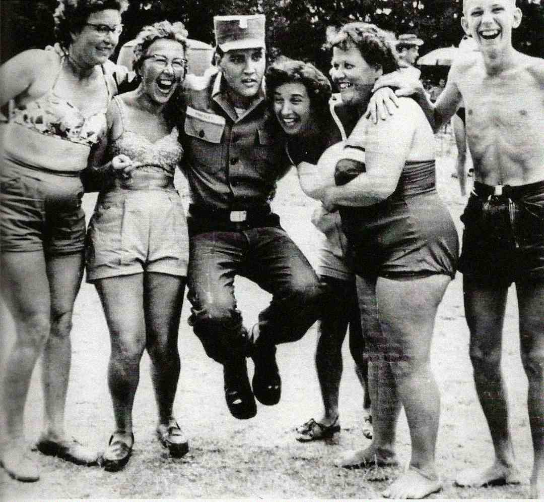 A man in a military uniform is playfully lifted off the ground by five smiling women in swimsuits, with another shirtless man standing beside them, outdoors on a sunny day.