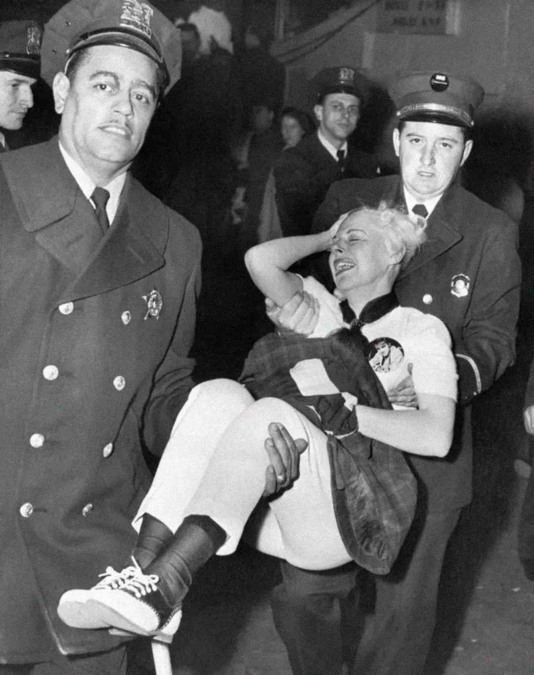 Two uniformed officers carry a woman who appears distressed or possibly injured; she is holding her head and has her eyes closed while other people in uniforms look on in the background.