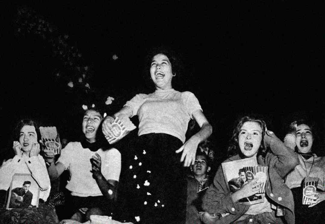 A group of excited teenage girls scream and cheer at a concert, holding snacks and magazines with a performer’s photo. The black-and-white image captures their energetic reactions and enthusiasm.