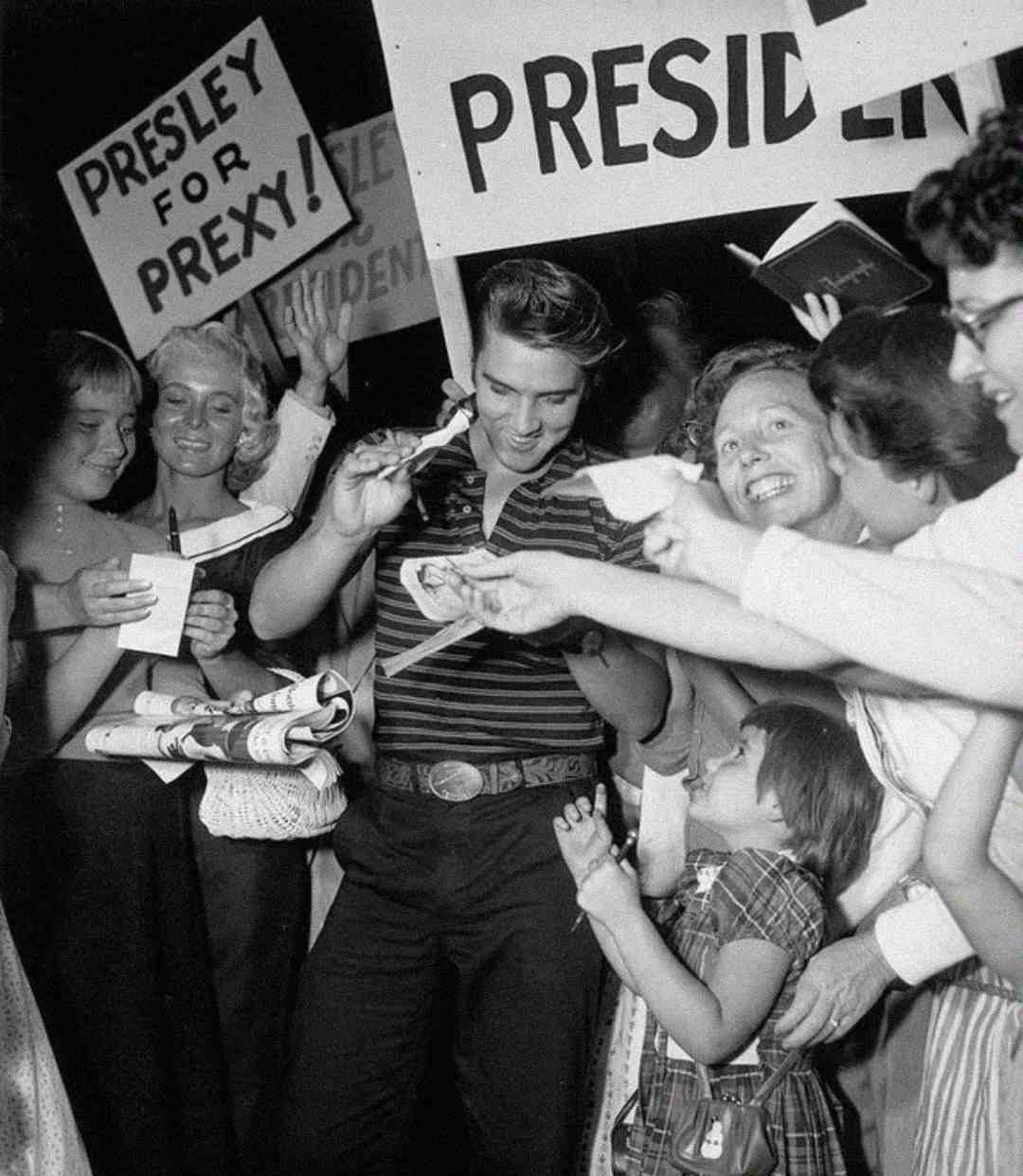 Elvis Presley smiles and signs autographs for an excited crowd of fans, some holding signs that read "PRESLEY FOR PREXY" and "PRESIDENT," in a black and white photo.