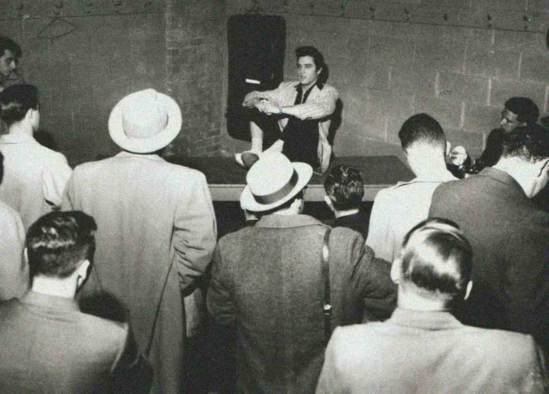 A young man sits on a stage or raised platform, knees up, surrounded by a crowd of people in suits and hats, all facing him in what appears to be a vintage black-and-white photo.