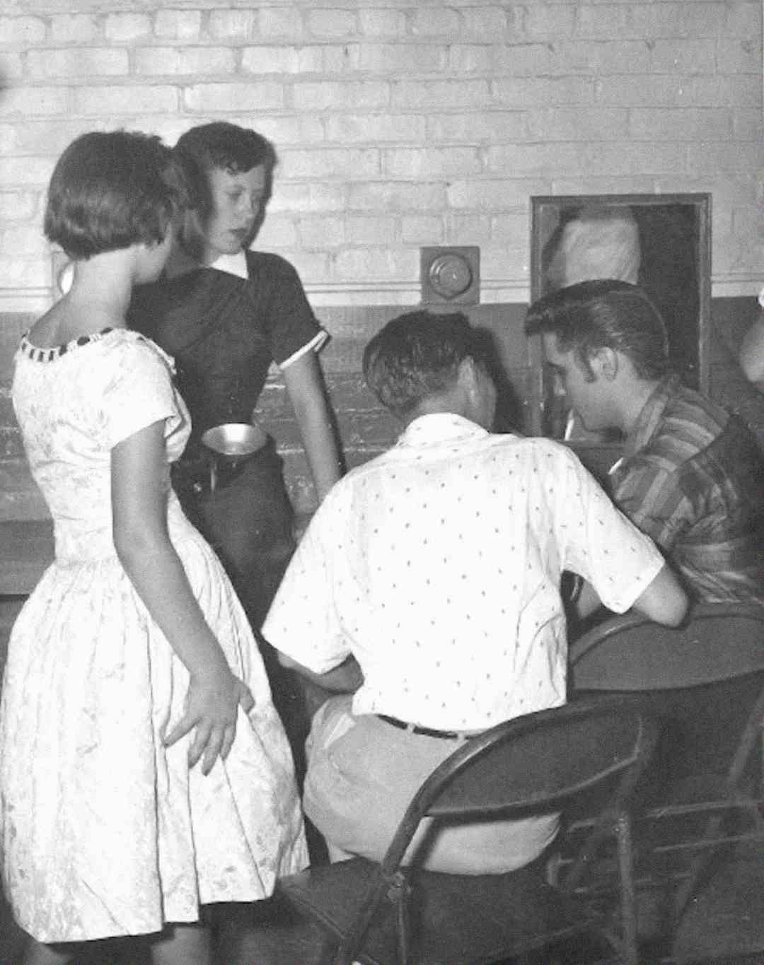 Two young women stand near two seated men, all dressed in 1950s-style clothing, in a room with brick walls and a small mirror in the background. The scene appears casual and conversational.