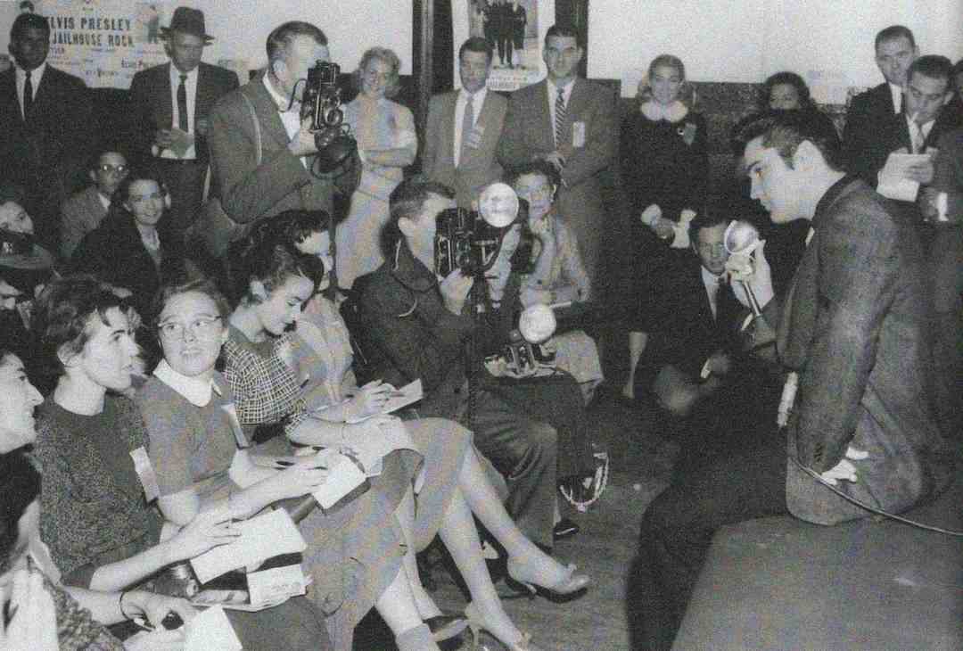 A young man sits and speaks into a microphone while a crowd of reporters, photographers, and onlookers watch and take notes and pictures in a busy room. A poster on the wall mentions "Elvis Presley." This is a black-and-white photo.