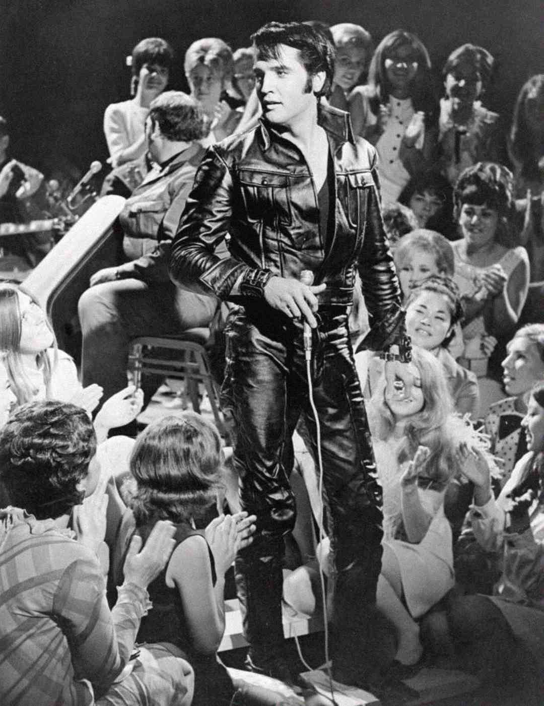 A man in a black leather outfit stands on stage holding a microphone, surrounded by an audience clapping and smiling. A band plays in the background. The image is in black and white.