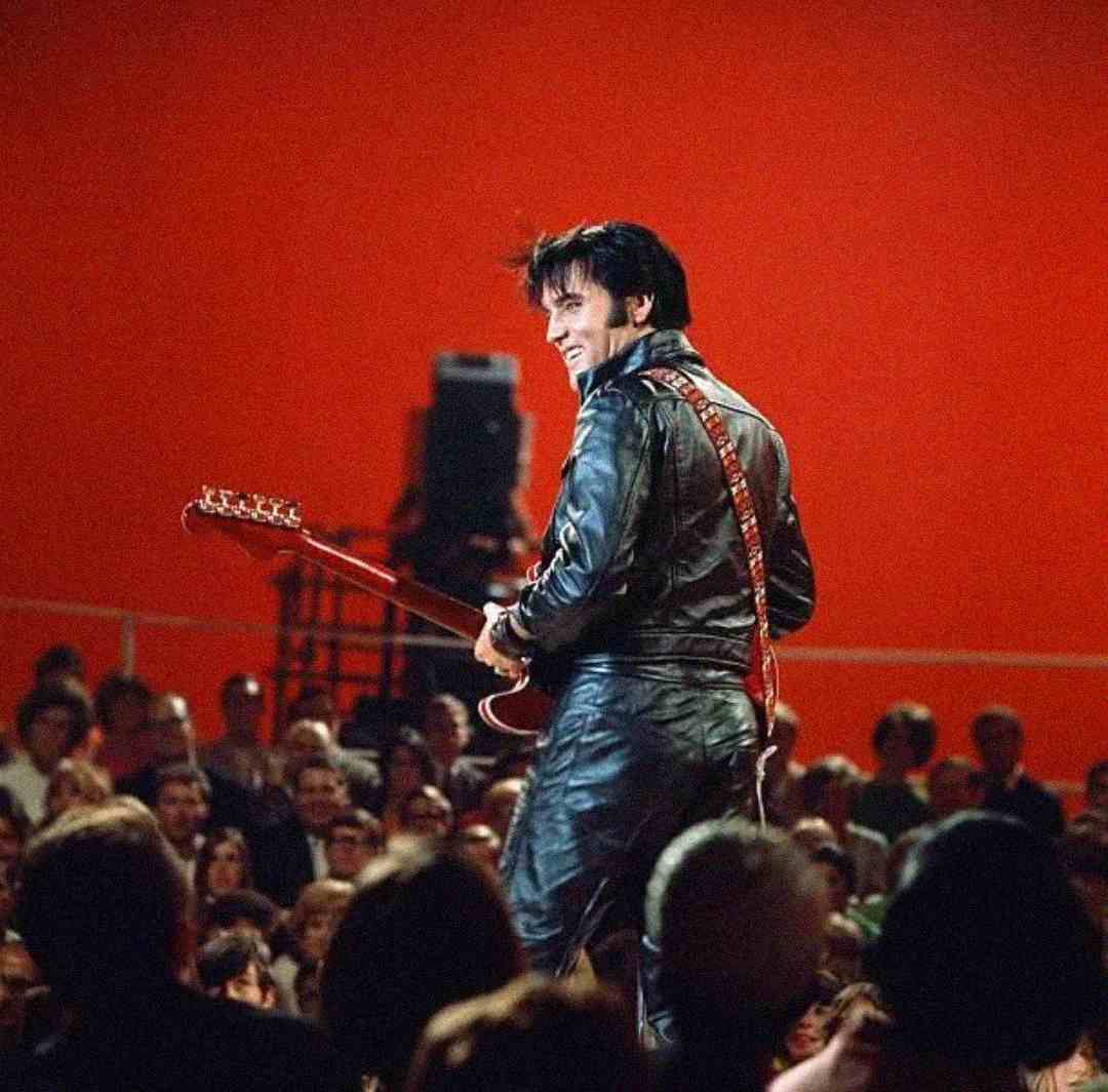 A man in a black leather outfit plays guitar on stage, smiling as he faces an audience. The background is bright red, and the crowd watches attentively.