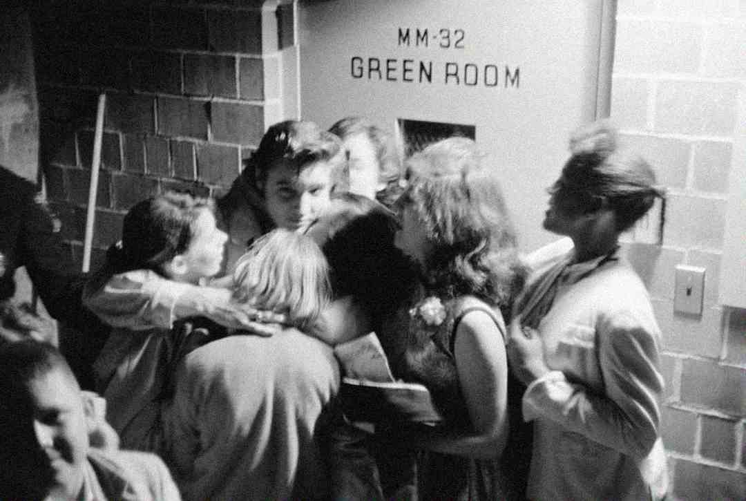 A group of young fans excitedly crowd around a man as he exits a door labeled "GREEN ROOM" in a brick hallway. The scene is lively and energetic, with fans reaching out to him.