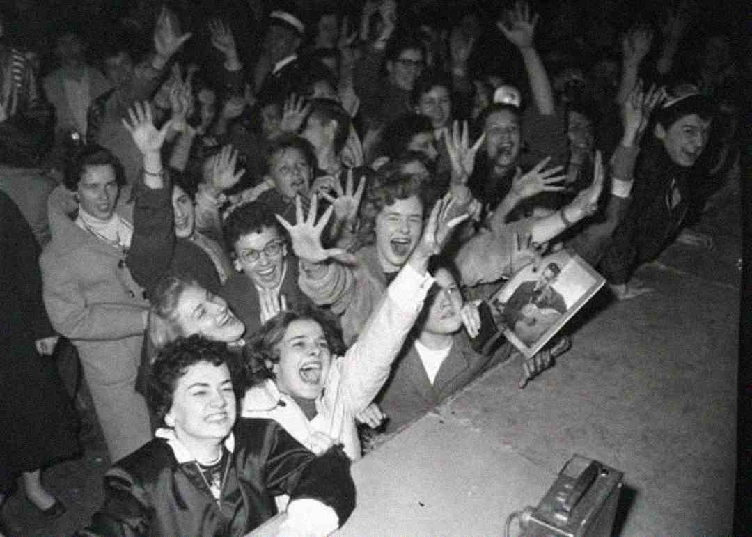 Black and white photo of a crowd of excited young women at a concert, smiling and waving their hands in the air. One holds a magazine or poster with a photo of a performer.