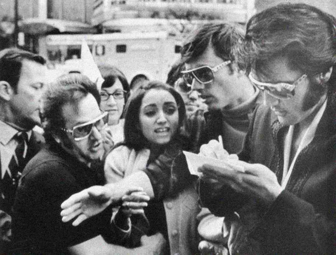 A black-and-white photo shows a woman surrounded by enthusiastic men in sunglasses; one man is writing on paper, while another gently holds the woman’s arm. Several onlookers watch in the background.