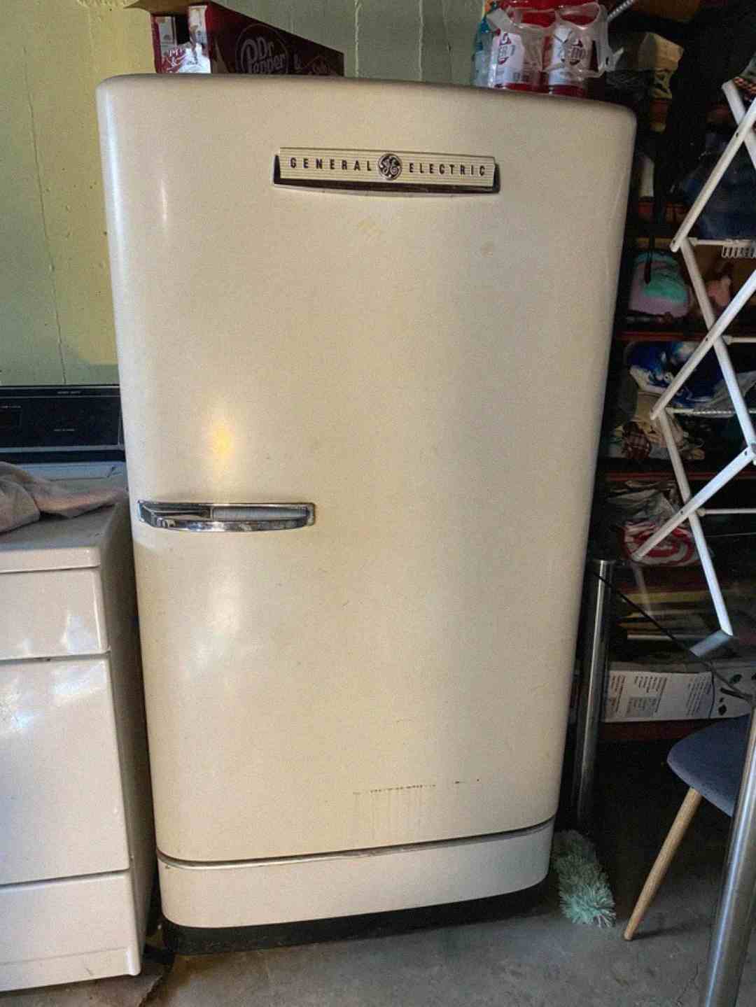 A vintage cream-colored General Electric refrigerator stands in a cluttered room, surrounded by a washing machine, shelves, and household items.