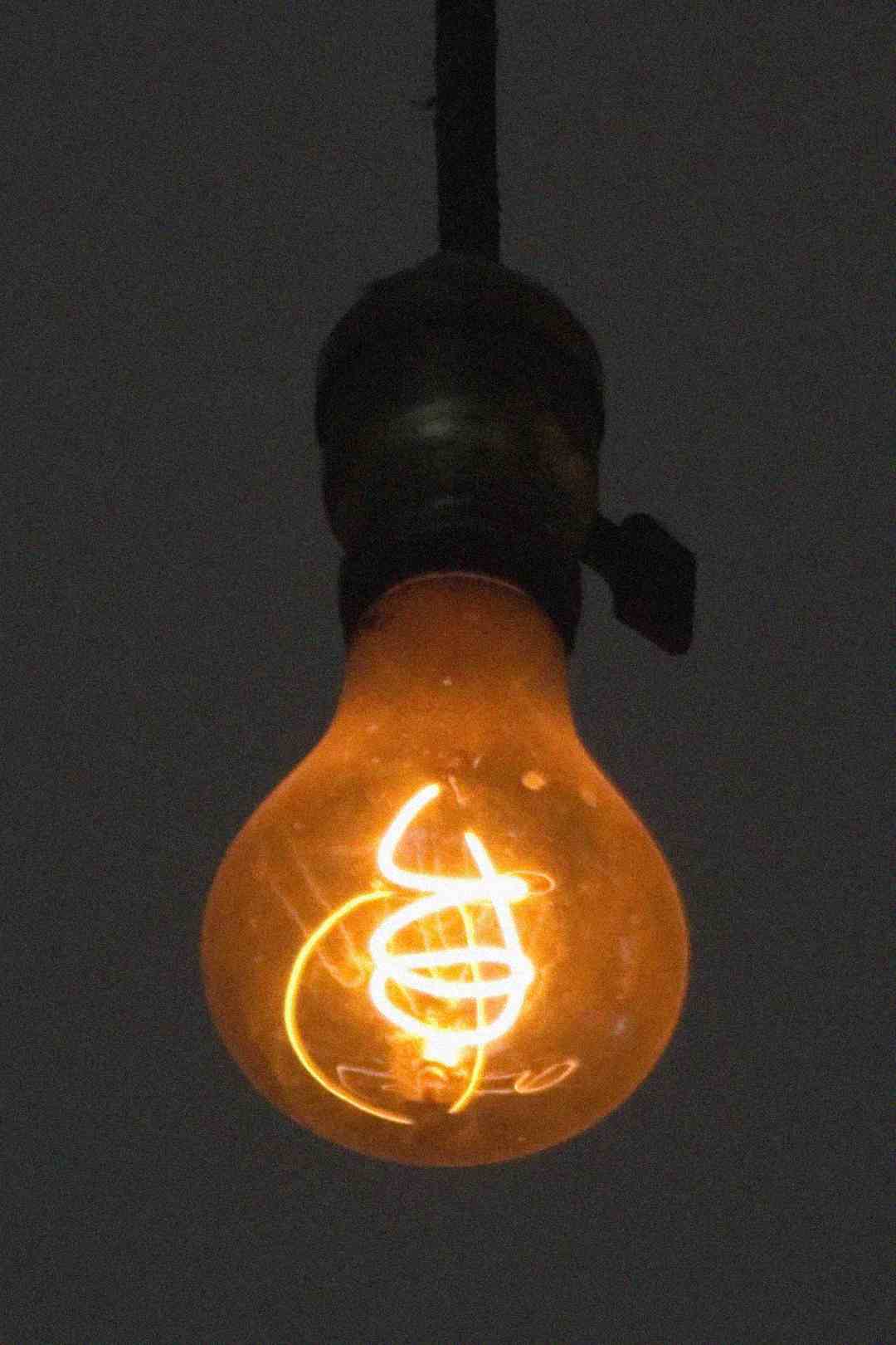 A single glowing incandescent light bulb hangs from a wire against a dark background, with its filament visibly illuminated in a warm orange hue.